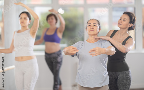Fotografie Focused female ballet choreographer patiently instructing senior woman in basic ballet techniques at group dance class for amateurs in spacious light-filled studio