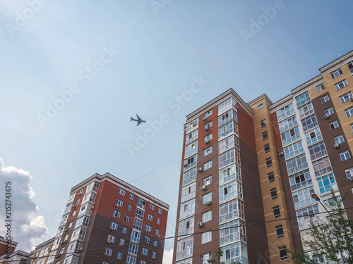 Airplane flying above tall residential apartment buildings against clear blue sky, low angle view, urban aviation concept for real estate, transportation, travel