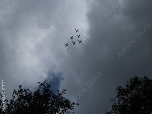 Several small aircraft fly together in formation across a cloudy sky above the treetops of a rural area in Java, Indonesia.
