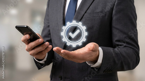 Businessman in a suit holding a smartphone and showing a digital quality check mark icon
