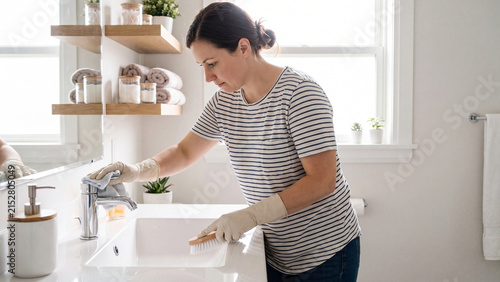 Woman wearing rubber gloves scrubs a modern white bathroom sink and faucet with a cleaning cloth during routine domestic housework.