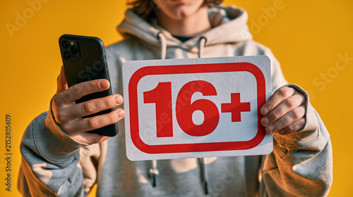 Age Limit 16+: A person holds a smartphone and a sign displaying 16+ against a vibrant yellow background, signifying an age restriction.