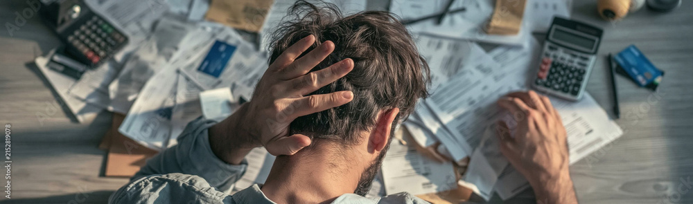 Fototapeta premium Overwhelmed by Debt: A man clutches his head in despair, surrounded by a chaotic pile of bills, receipts, and calculators, symbolizing financial stress and overwhelming debt.