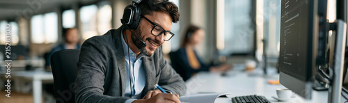 Dedicated Call Center Agent: A focused male customer service representative wearing a headset, eyeglasses, and a suit, works diligently at his desk in a modern office, assisting clients with a smile.