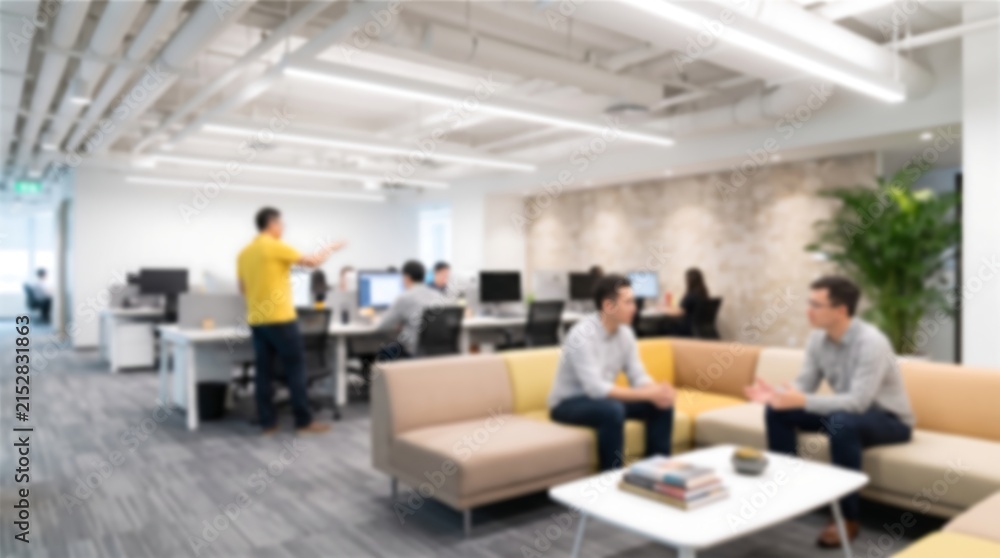 Fototapeta premium Blurred open plan corporate office space with employees sitting on sofa in lounge area having meeting while team members work at desks in background under bright linear lighting