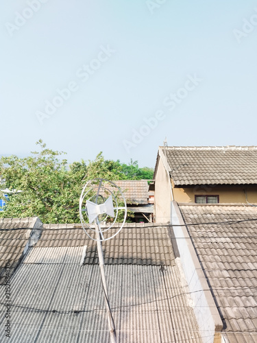 Clear sky above village rooftops and green trees. Metal antenna stands tall on a tiled roof. Houses blend into distant hills and calm air. Scene evokes quiet daily life in rural setting