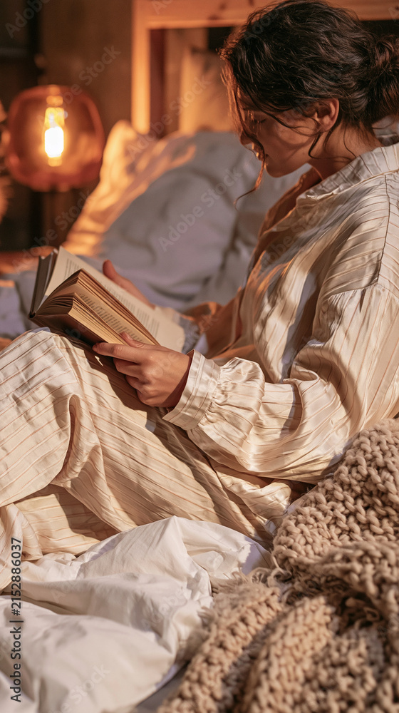 Obraz premium Cozy Evening Read: A young woman in comfortable sleepwear relaxes in bed, engrossed in a book under the warm glow of a bedside lamp.