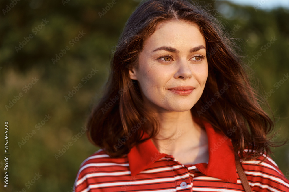 Obraz premium A smiling young woman in a striped shirt enjoys a sunny day at the park. Her lifestyle portrait shows natural beauty, outdoor leisure, happiness, and authentic joy.