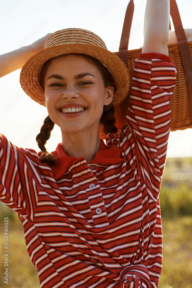 Obraz premium A smiling young woman in a striped shirt and straw hat carries a picnic basket outdoors. Lifestyle concept for summer leisure, joy, relaxation, and nature travel.