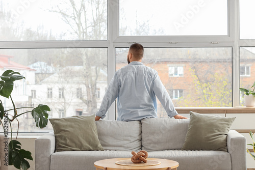 Young man behind sofa looking out large window at home, back view