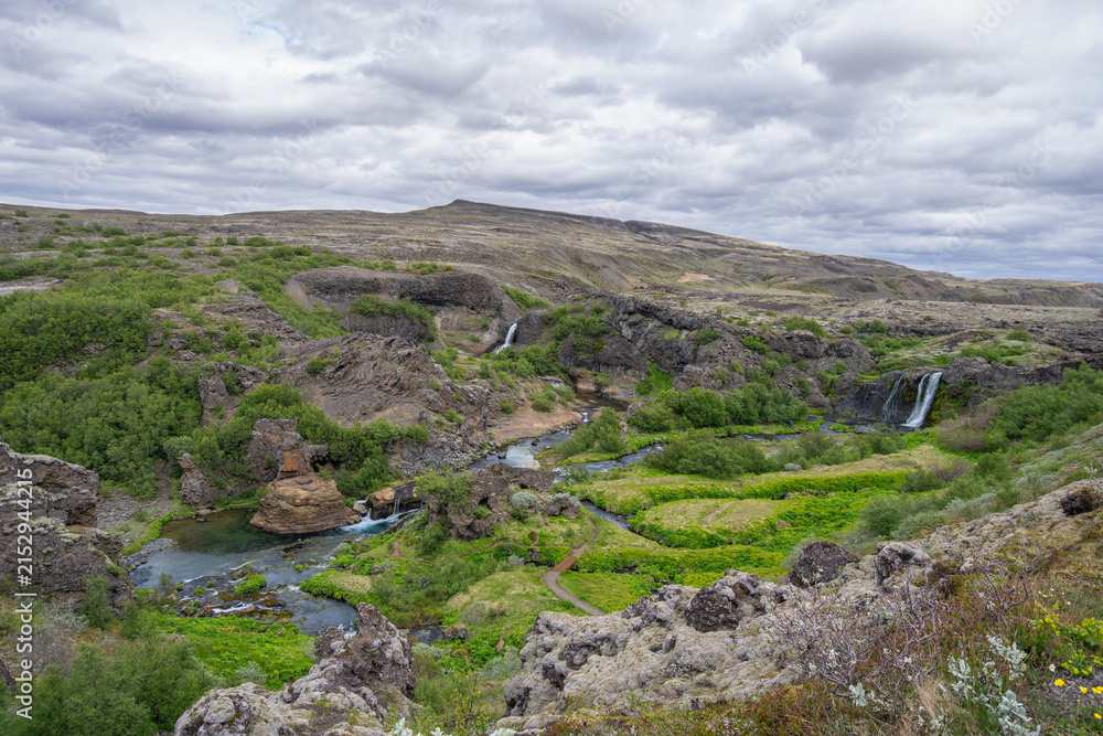 Fototapeta premium Scenic landscape of the lush green Gjain valley in Thjorsardalur, Iceland, featuring the Rauda river, small waterfalls, and volcanic rock formations.