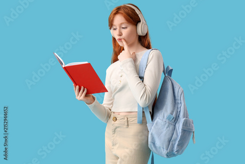 Thoughtful teenage redhead girl with notebook and backpack on blue background