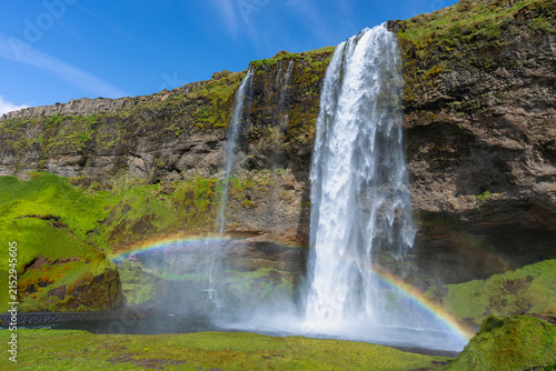 Scenic view of the famous Seljalandsfoss waterfall cascading down green cliffs with a vibrant rainbow in Iceland.