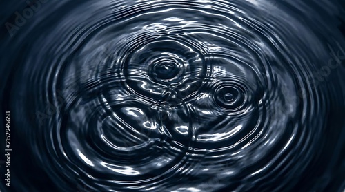 Close-up view of intersecting circular ripples on a dark, reflective water surface, showing the fluid motion and concentric wave patterns in deep tones