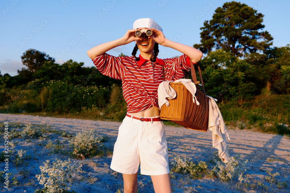 Obraz premium Young woman with binoculars looking ahead at sunset. Picnic basket and stylish outfit, summer lifestyle outdoors. Happy girl exploring nature in a bright sunny landscape.