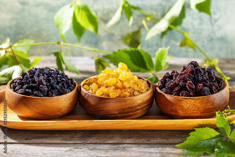 Fototapeta premium Close up raisin assortment with golden, black and dark red raisins in wooden bowls on rustic table with grape leaves