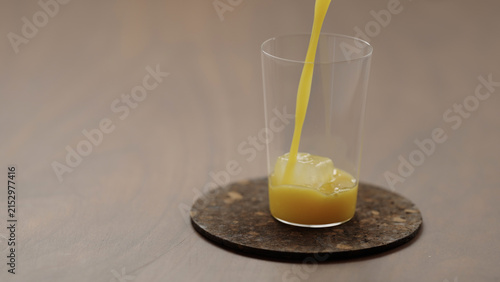 Konstfotografi pour orange juice in a glass with ice cube on a wood table