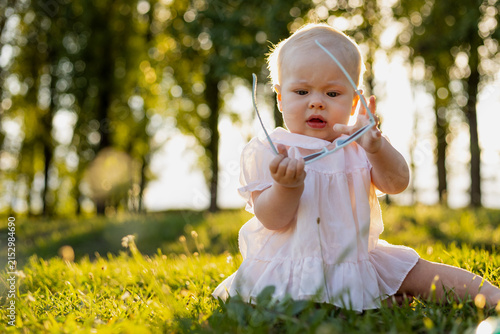 Curious baby girl in a pink dress explores sunglasses outdoors in golden hour light