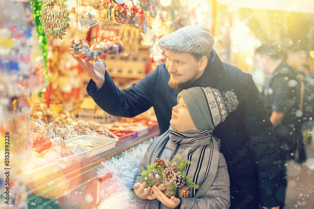 Obraz premium Man with son among crowd of shoppers at holiday fair. Buyers by outdoor souvenir shop window examines Christmas toys and interior decorations, choose balls and Christmas tree toys in window of fair.