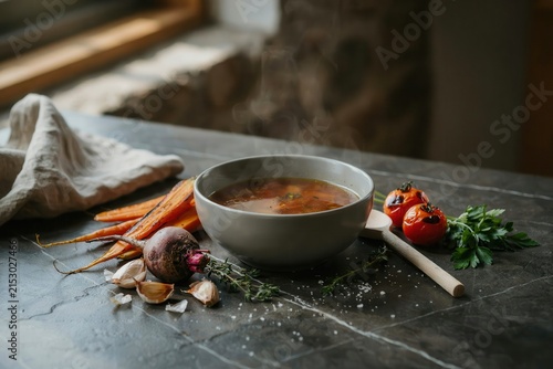 Hot vegetable soup in a ceramic bowl with roasted tomatoes, carrots, beetroot, garlic, and fresh herbs on a dark grey marble surface