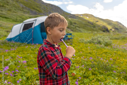 Child, cute boy, brushing his teeth in front of a tent in the morning in a beautiful Norway nature, grass, mountains, flowers, anazing nature