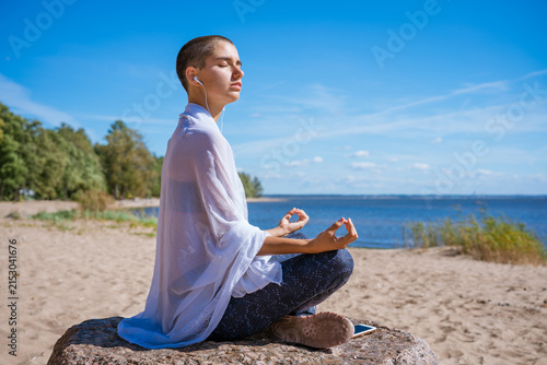 Quadro su tela Woman wrapped in a white shawl sits in a lotus pose on a stone by the sea, a spa ritual, meditation and coastal wellness concept in summer daylight