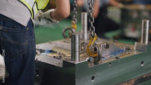 Worker lifts heavy metal mold with chain hook. Industrial setting, focused on manufacturing process. Equipment shows signs of use and wear