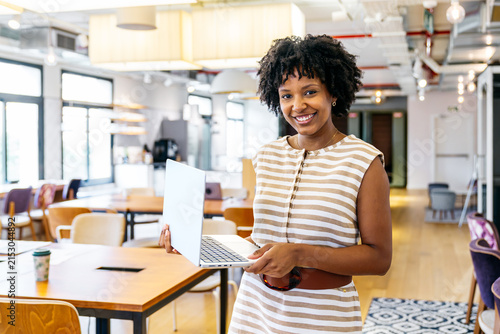 Young black businesswoman smiling, holding a laptop, working remotely in a contemporary shared office space with natural light