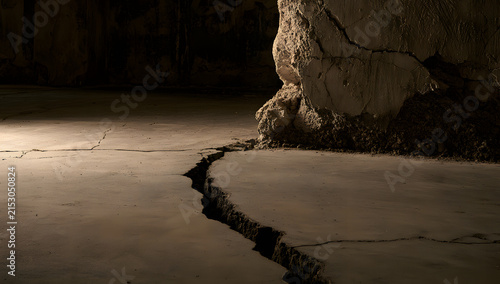 Concrete flooring with extensive splits and a shattered cement wall in a poorly lit space, revealing signs of earthquake damage and structural weakness.