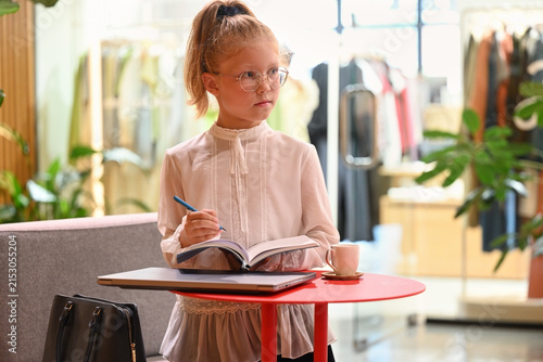 A little girl stands at a red table with a pen and a notebook in her hands