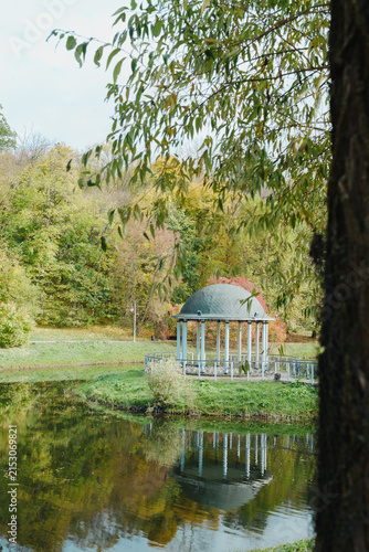 Fotografía Charming gazebo surrounded by trees reflects in a serene lake under a blue sky