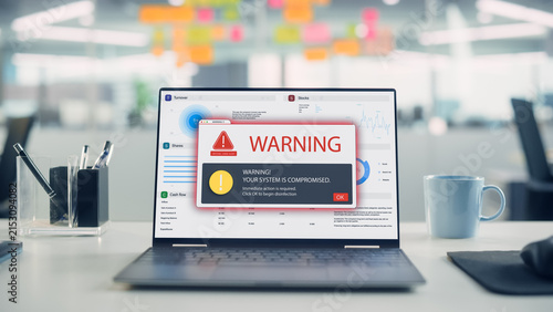 Laptop on Desk Displaying a Critical Virus Alert Warning Pop-up Over Financial Charts, Signaling a Compromised System in Open Plan Office Interior.