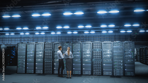 Two Caucasian IT Engineers, Stand With Their Backs Turned Inspecting Server Rack Cabinets in a Vast Modern Data Center Illuminated by Rows of Bright Ceiling Lights