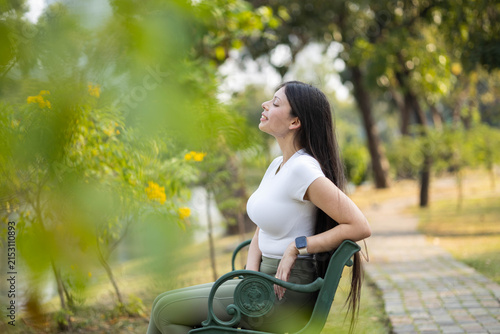 Young woman sitting on a park bench, eyes closed, enjoying the warm sunshine and fresh air, feeling serene and peaceful
