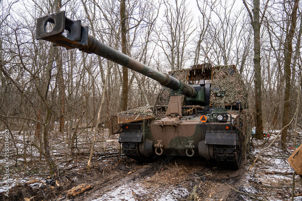 Obraz premium AHS Krab self-propelled howitzer of the Ukrainian Armed Forces camouflaged with a camouflage net and positioned in a winter forest. 