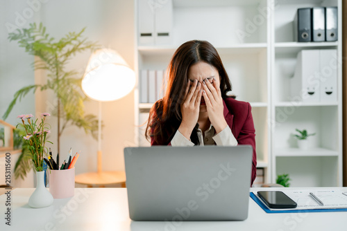Young asian businesswoman sitting at her desk. Covering her face with hands. Expressing severe stress. Headache. Or burnout while working on a laptop in a modern office
