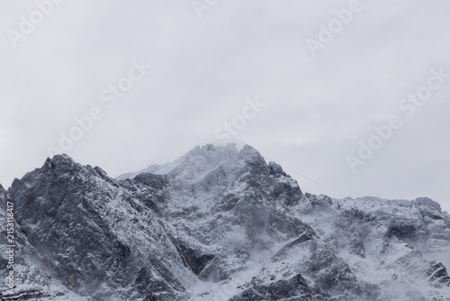 Majestätischer, schneebedeckter Berggipfel in den Alpen, umgeben von dichten Wolken und Nebelschwaden. Dramatische Winterlandschaft mit Felsformationen bei bewölktem Himmel.