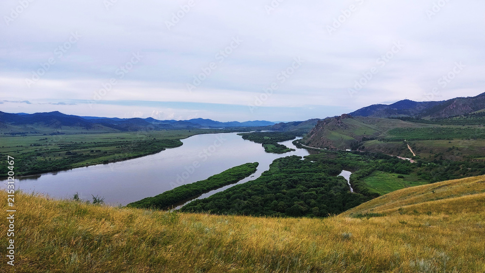 Fototapeta premium Winding Selenga River flowing through lush green valleys and forested islands in Buryatia, Siberia, with rolling hills under an overcast sky