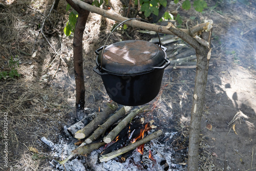A black cast iron Dutch oven pot with a lid hangs over an active outdoor campfire with burning wood and embers, suspended from a rustic wooden tripod structure in a forest for wilderness cooking.