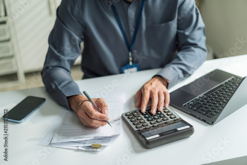 Close-up of office worker holding pen and using calculator to check financial bills and documents at desk.