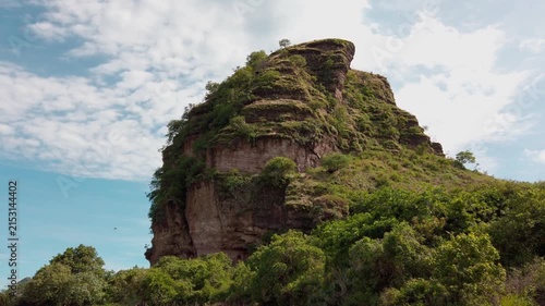 Scenic view of a massive layered rock formation covered with lush vegetation, rising above dense greenery beneath a blue sky with scattered clouds in Mexico
