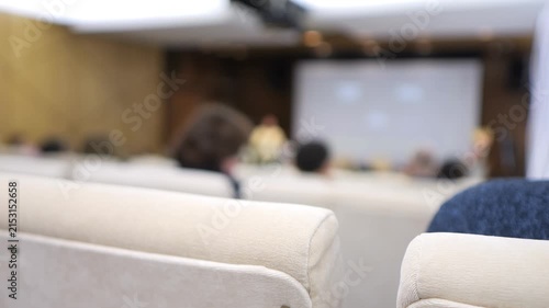 Blurred conference room with beige chairs and screen. People sit, facing forward, focused on presentation. Ideal for business, training, or educational video clips