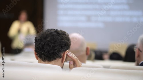 Audience listens attentively during a business presentation. Man in the front row touches his ear. Several attendees focus on the speaker. Projector screen displays data behind the presenter