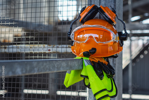 Orange hard hat with safety goggles, ear defenders, work gloves and reflective vest hanging on metal beam at construction site. Occupational health and safety equipment concept for industrial.