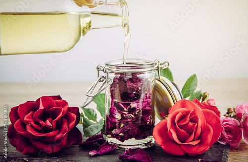 A person pours carrier oil over dried rose petals in a jar to make rose-infused oil at home. Rose blossoms in different colors arranged decoratively around it.