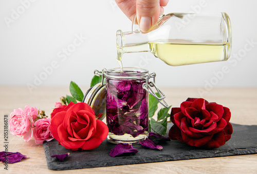 A person pours carrier oil over dried rose petals in a jar to make rose-infused oil at home. Rose blossoms in different colors arranged decoratively around it.