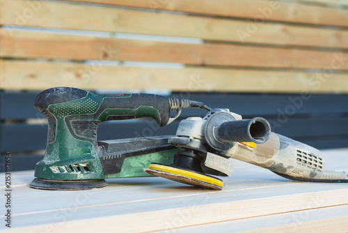 Electric orbital sander and angle grinder with sanding disc on wooden planks outdoors. Woodworking power tools, carpentry equipment or surface polishing concept for hardware store website banner.