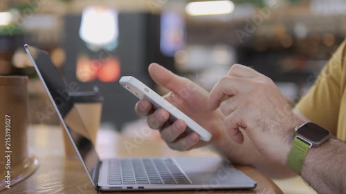 Person multitasks using phone and laptop at cafe. Hand holds phone, taps screen. Wears smartwatch, yellow shirt. Focuses on work or social media. Suitable for tech, productivity