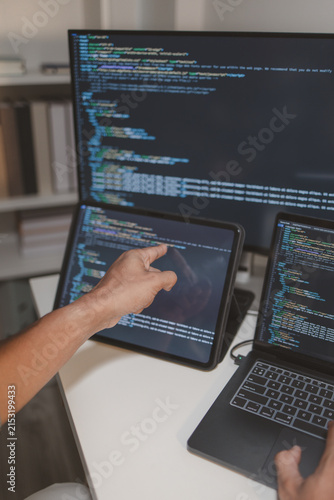 Professional software developer coding across laptop, tablet, and monitor in a modern workspace. Multi-device programming, debugging, software engineering, and application development workflow.