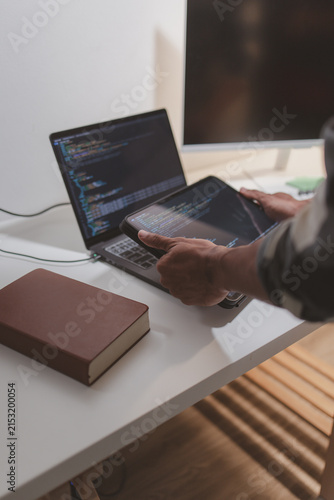 Software developer coding on laptop and tablet in a modern workspace. Close-up of programming workflow, software engineering, debugging source code, and multi-device application development.